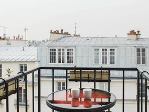 Un balcon avec une table et deux tasses, donnant sur une rangée d’immeubles dotés de nombreuses cheminées.