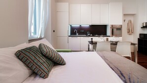 Ein modernes Hotelzimmer mit Bett, Kissen, einem kleinen Tisch mit Stuhl, einer Küchenzeile und einem Fenster mit Vorhängen.