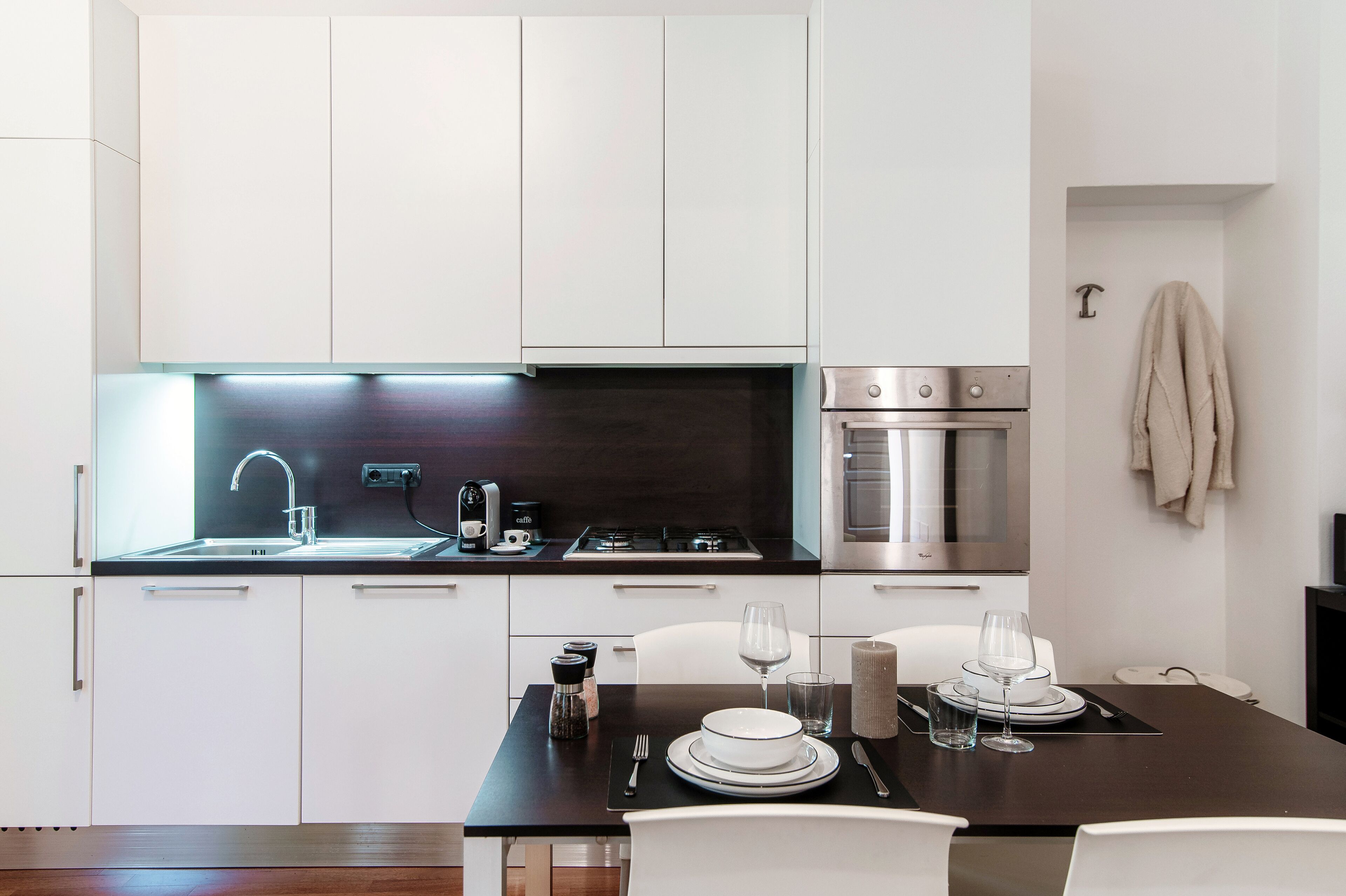 A modern kitchen with white cabinets, a dark countertop, a gas stove, an oven, and a dining area with a table set for two.