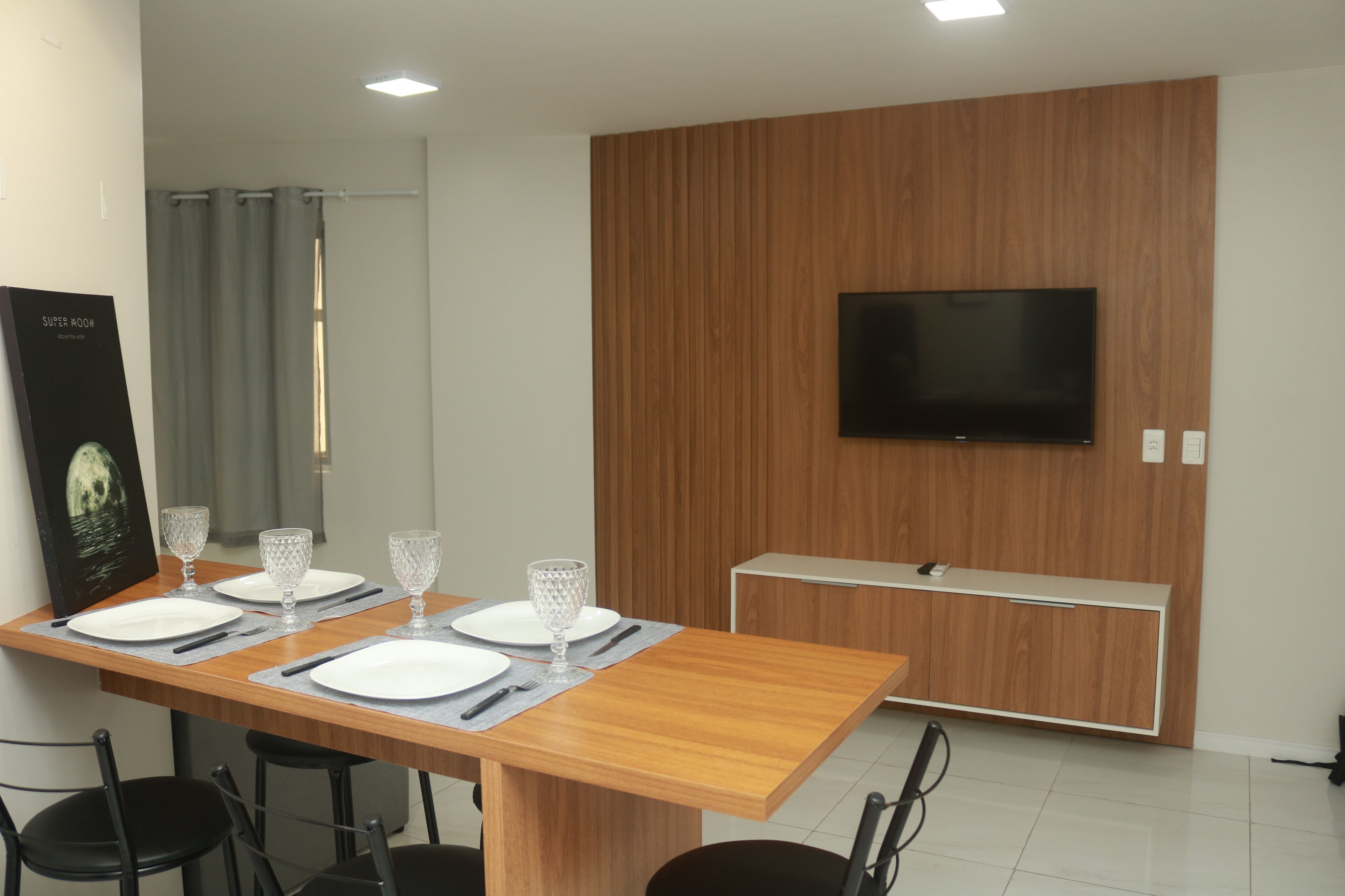 Un comedor moderno con una mesa de madera dispuesta para cuatro personas, un televisor de pantalla plana montado en una pared de paneles de madera, y un cartel a la izquierda.