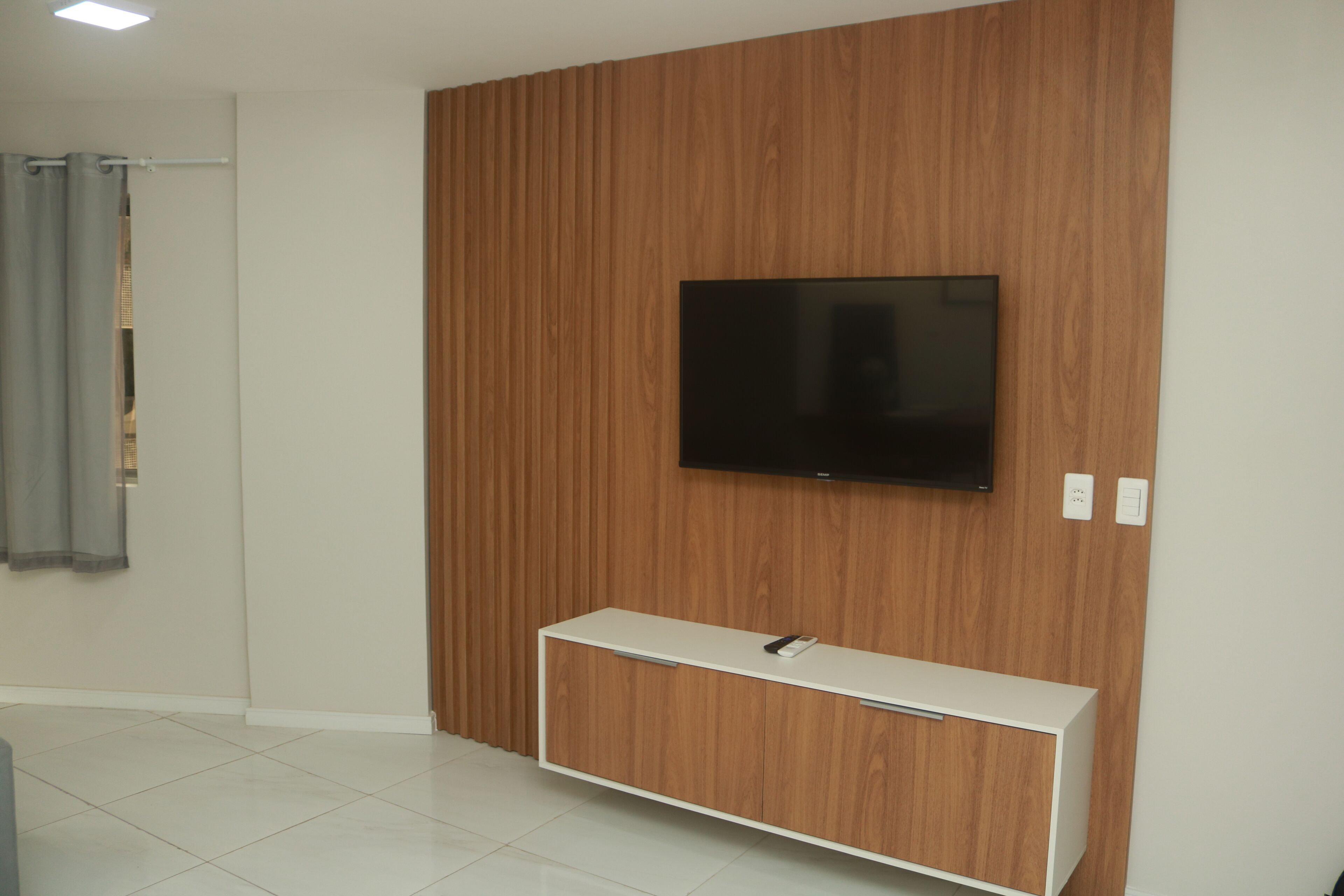 A modern living room with a wooden panel wall, a flat-screen TV mounted above a white console, and a grey curtain on the left.