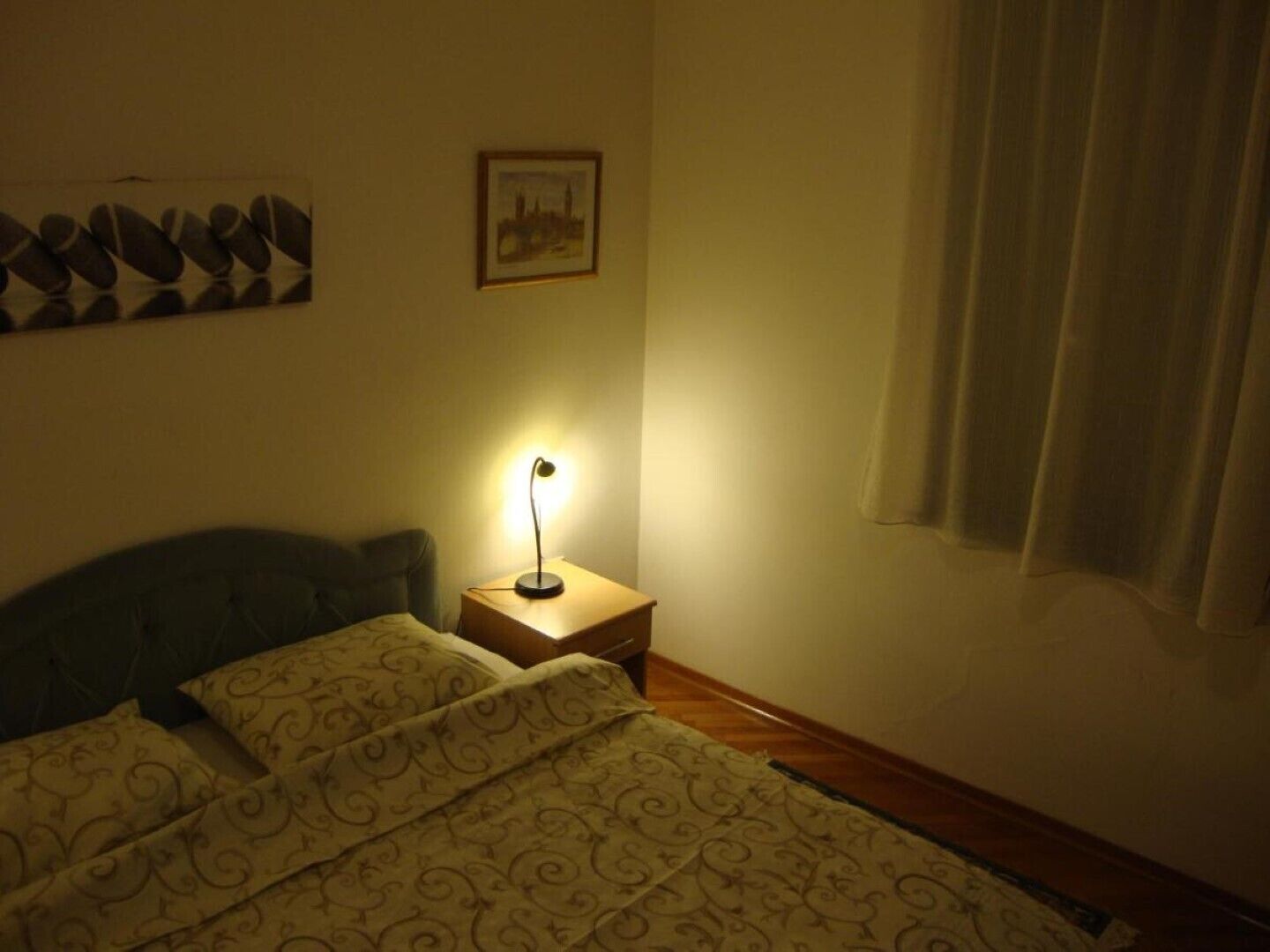 Quarto com cama, mesa de cabeceira com abajur, quadro na parede e janela com cortinas.
