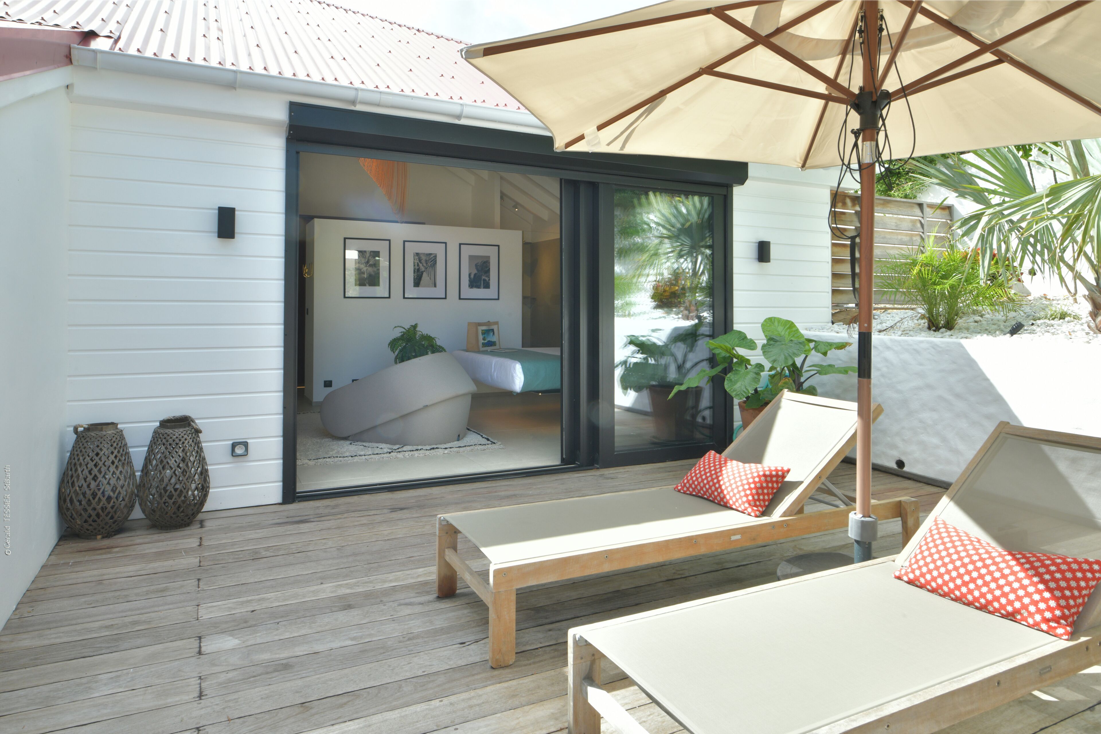 A wooden deck with two lounge chairs and an umbrella, a sliding glass door leading to a bedroom with a bed and a potted plant.