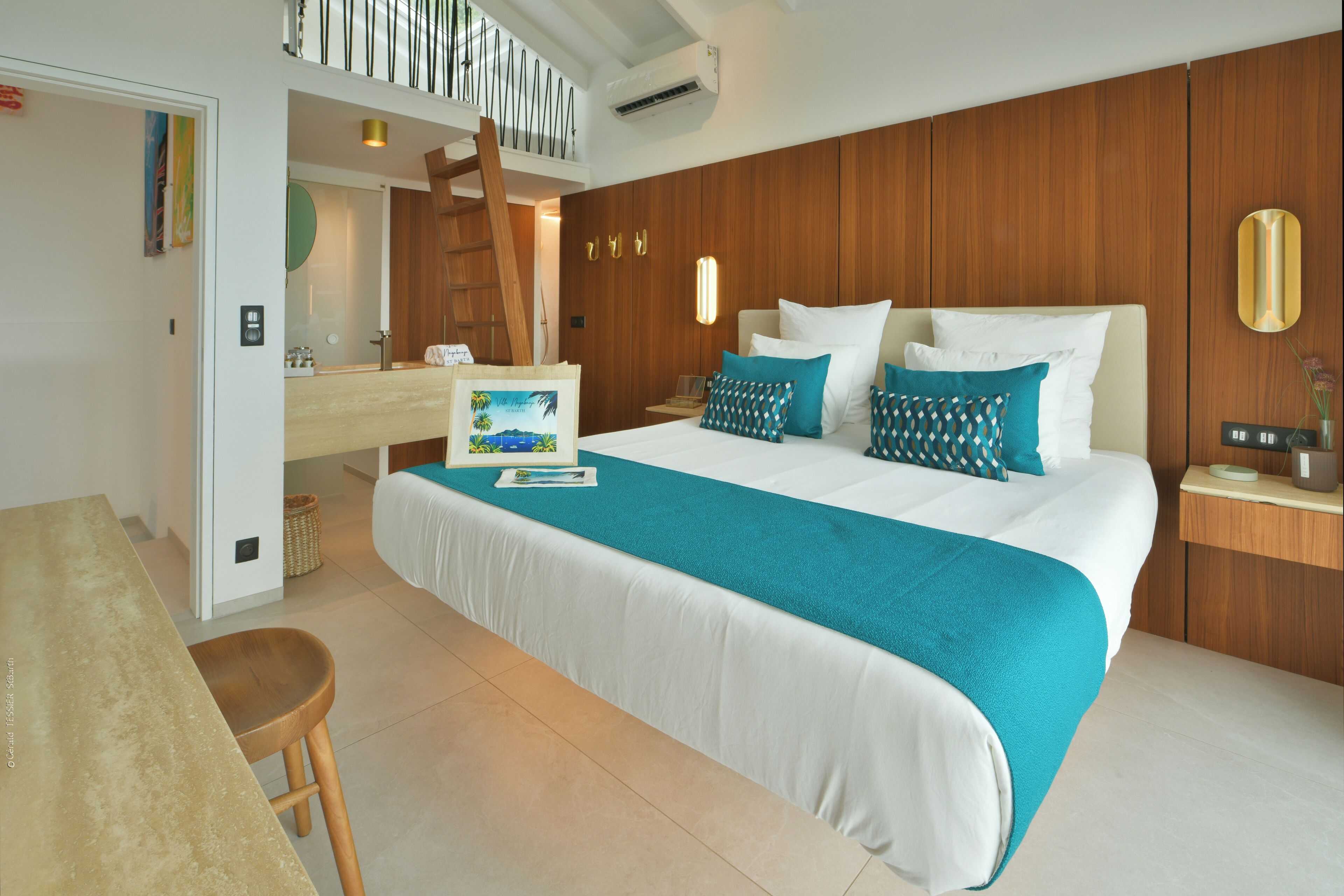 Quarto de hotel com uma cama grande, cabeceira de madeira, mesa de cabeceira com abajur e um banheiro visível através de uma porta aberta.