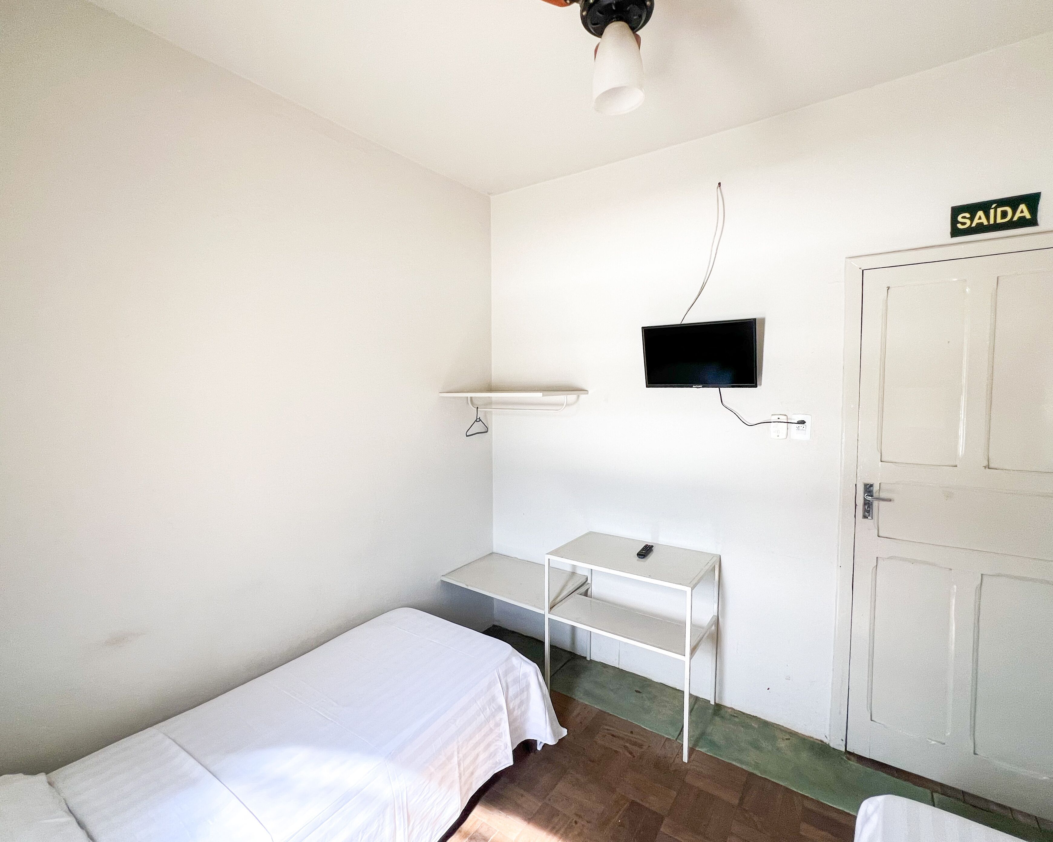 Una habitación pequeña y blanca con una cama, un escritorio, un televisor montado en la pared y una puerta con la etiqueta “SAÍDA”.