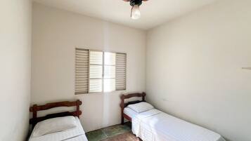 Une petite chambre propre, avec deux lits simples, un ventilateur de plafond et une fenêtre avec des volets.
