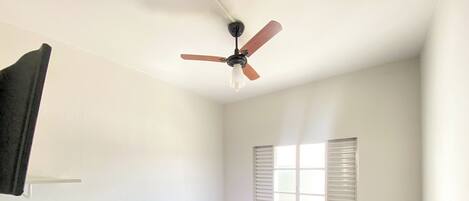 Une petite chambre avec un lit simple, un ventilateur de plafond, une fenêtre avec des stores et un tapis vert sur le sol.