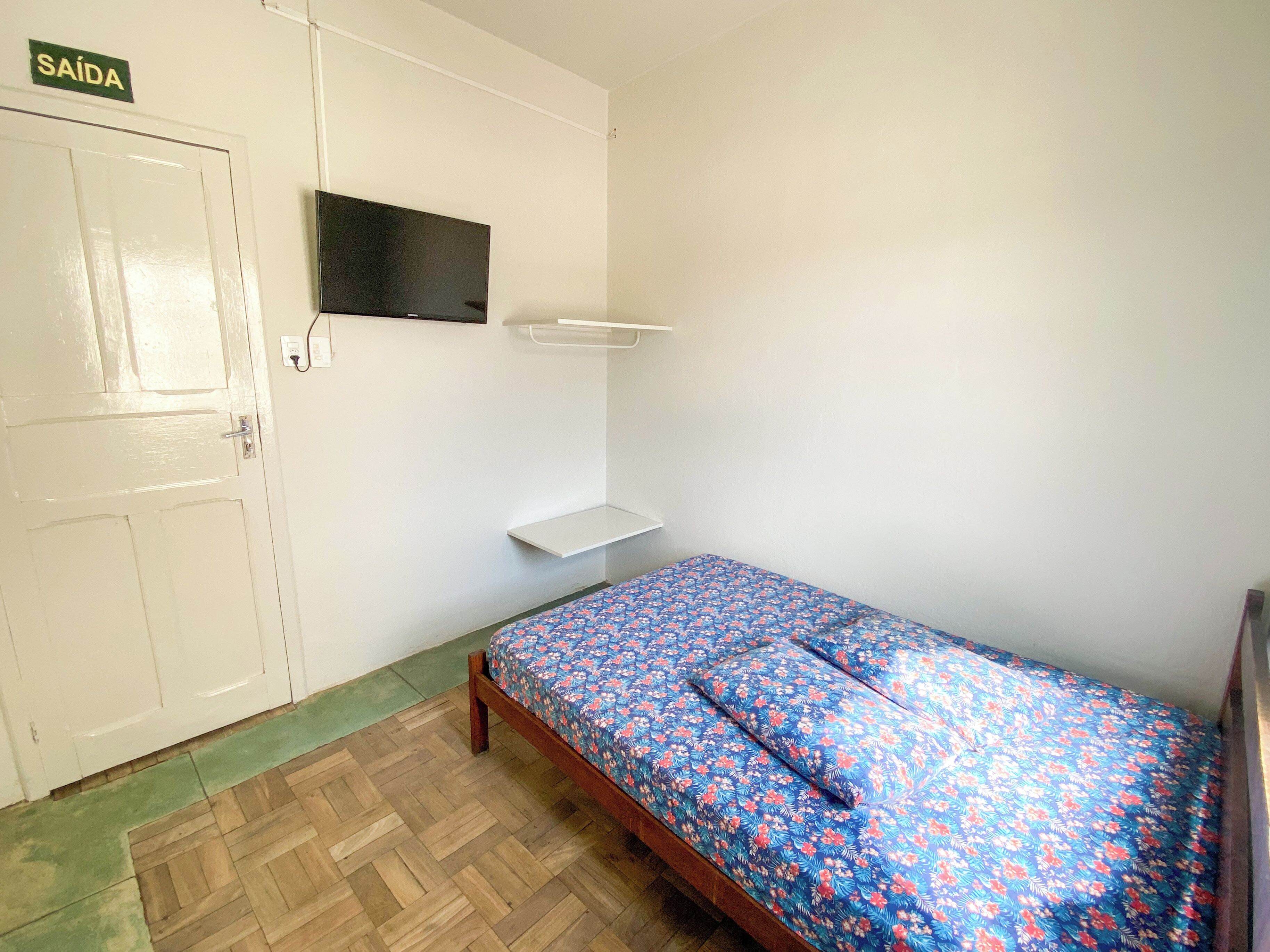 Una habitación pequeña con cama individual, piso de madera, ropa de cama con estampado floral, una puerta blanca con un letrero encima y un televisor montado en la pared.