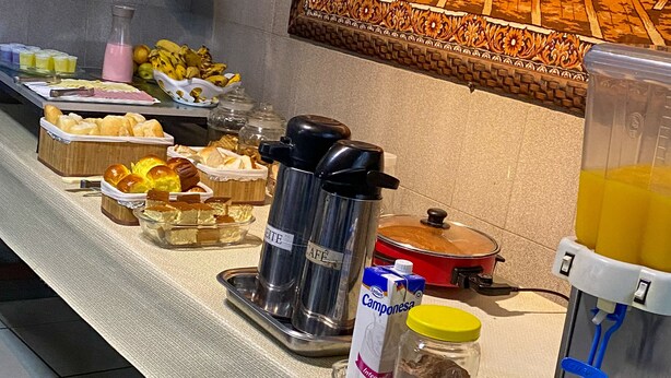 A buffet table with various food items, a coffee maker, a juice dispenser, and a painting on the wall. - Elite Economy Hotel (Para de Minas)