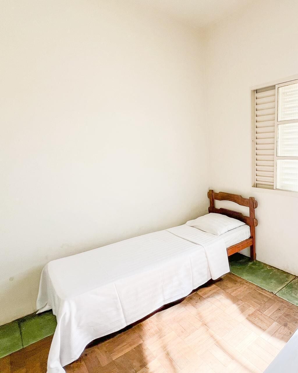 Una habitación pequeña con una cama individual, cabecera de madera, colcha blanca y una ventana con persianas.