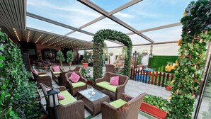 Un área de estar al aire libre techada con mobiliario de mimbre y cojines verdes, rodeada de plantas en macetas y un arco decorativo.