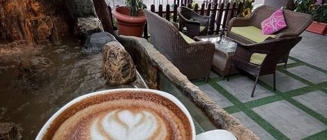 Una taza de café con arte de latte sobre una superficie de piedra en un área exterior con mobiliario de mimbre y una pared decorativa.