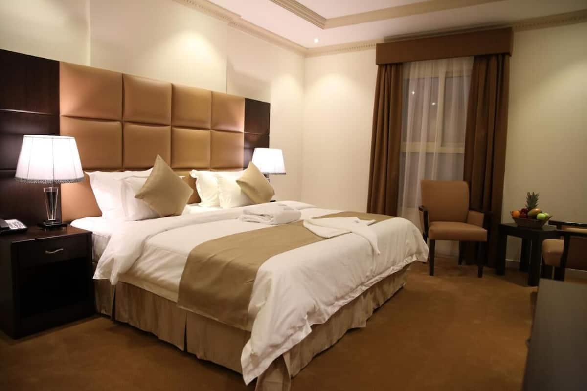 Une chambre d’hôtel avec un grand lit, deux tables de chevet avec des lampes, une chaise, une petite table avec des fruits et une fenêtre avec des rideaux.