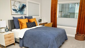 A bedroom with a bed, bedside tables, a nightstand, a lamp, a wall art piece, and a window with curtains. - Bright and sunny 1-bdr guest suite in South Calgary (De Winton)