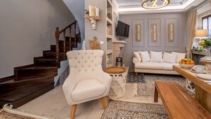 Una sala de estar con una escalera, un sillón blanco, un sofá, una mesa de centro de madera y un televisor montado en la pared.
