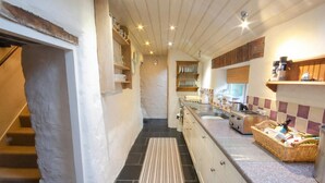A narrow kitchen with a central island, built-in shelves, and a tiled floor. - Tal y Gaer Cottage, Fishguard (Trefasser)