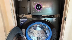 A compact, modern front-loading washing machine with a digital control panel, located in a small laundry room with wooden flooring.