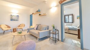 Ein modernes Wohnzimmer mit einer Couch, zwei Sesseln, einem Couchtisch und einem Beistelltisch mit einer Pflanze. Ein Durchgang führt in einen weiteren Raum mit einem Bett und einem Fenster.
