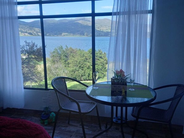 A room with a large window offering a view of a lake and mountains. There is a round glass table with two chairs and a bottle of Corona beer.