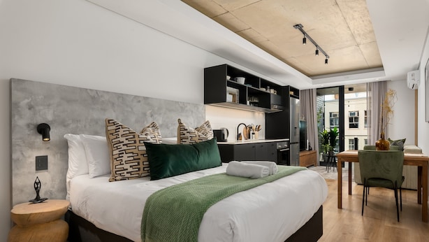 A modern bedroom with a large bed, a desk, and a kitchenette. - Station House Luxury Living - Generator + Pool (Cape Town)