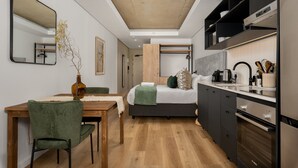 A compact, modern room with a wooden dining table, a green upholstered chair, a bed with a green throw, and a kitchenette with a built-in oven and microwave. - Station House Luxury Living - Generator + Pool (Cape Town)