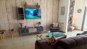 En stue med et stort fladskærms-tv, en sofa, et sofabord og et skænk med dekorative genstande.