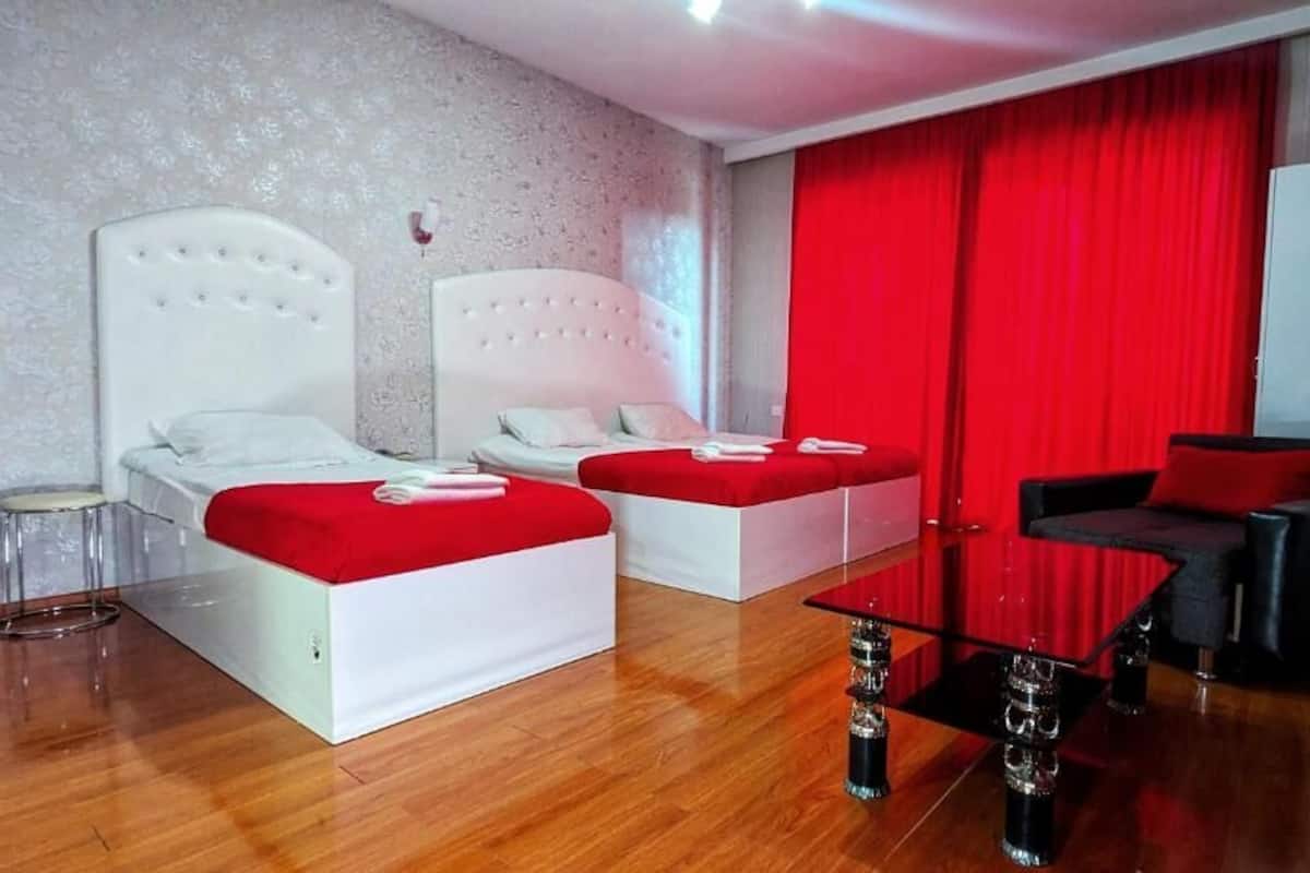 Habitación de hotel con dos camas, un sofá, una mesita de centro de cristal y cortinas rojas.