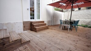 Une terrasse en bois avec une table et des chaises sous un parasol rouge, un ensemble de marches en bois menant à une porte, et un mur blanc avec une fenêtre.