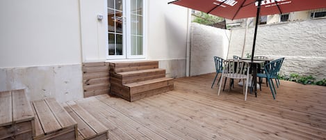 A wooden deck with a table and chairs under a red umbrella, a set of wooden steps leading to a door, and a white wall with a window.