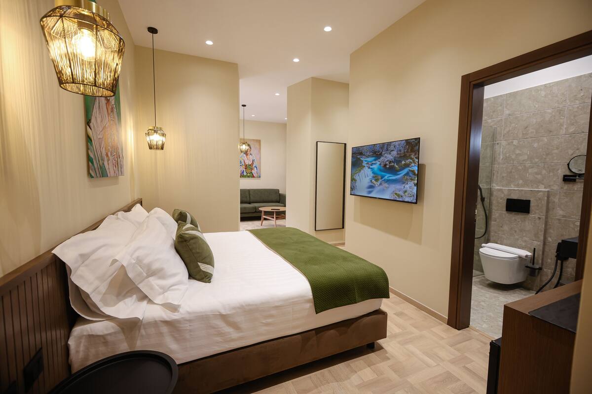 A modern bedroom with a large bed, a green throw blanket, and a wooden headboard. There is a painting on the wall and a ceiling light fixture.