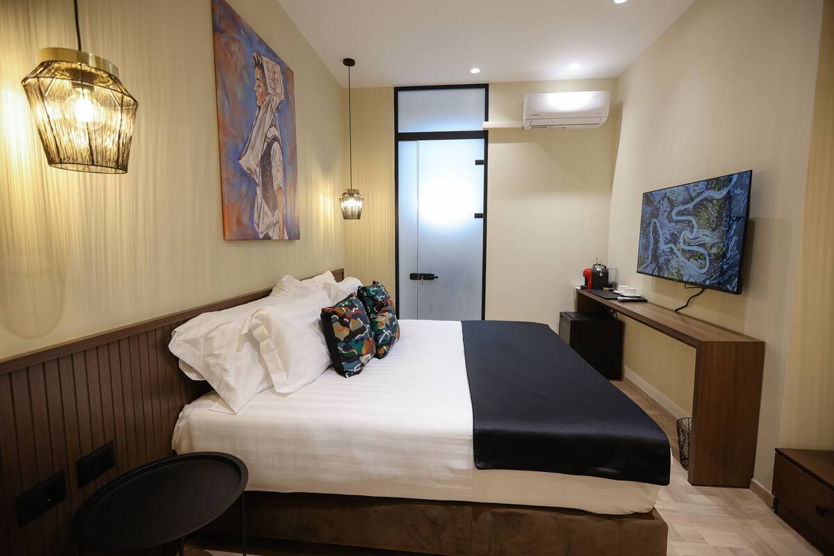 A hotel room with a bed, bedside table, chair, artwork, and a television.