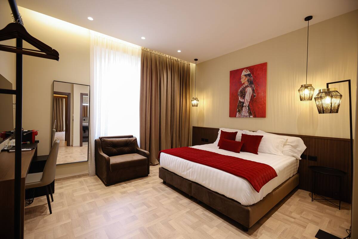 A modern hotel room with a large bed, a desk, a chair, and a wardrobe.