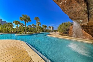 A swimming pool with clear blue water, surrounded by palm trees and a rocky cliff. - Elegant Destin Condo w/ Ocean Views: Walk to Beach (Destin)