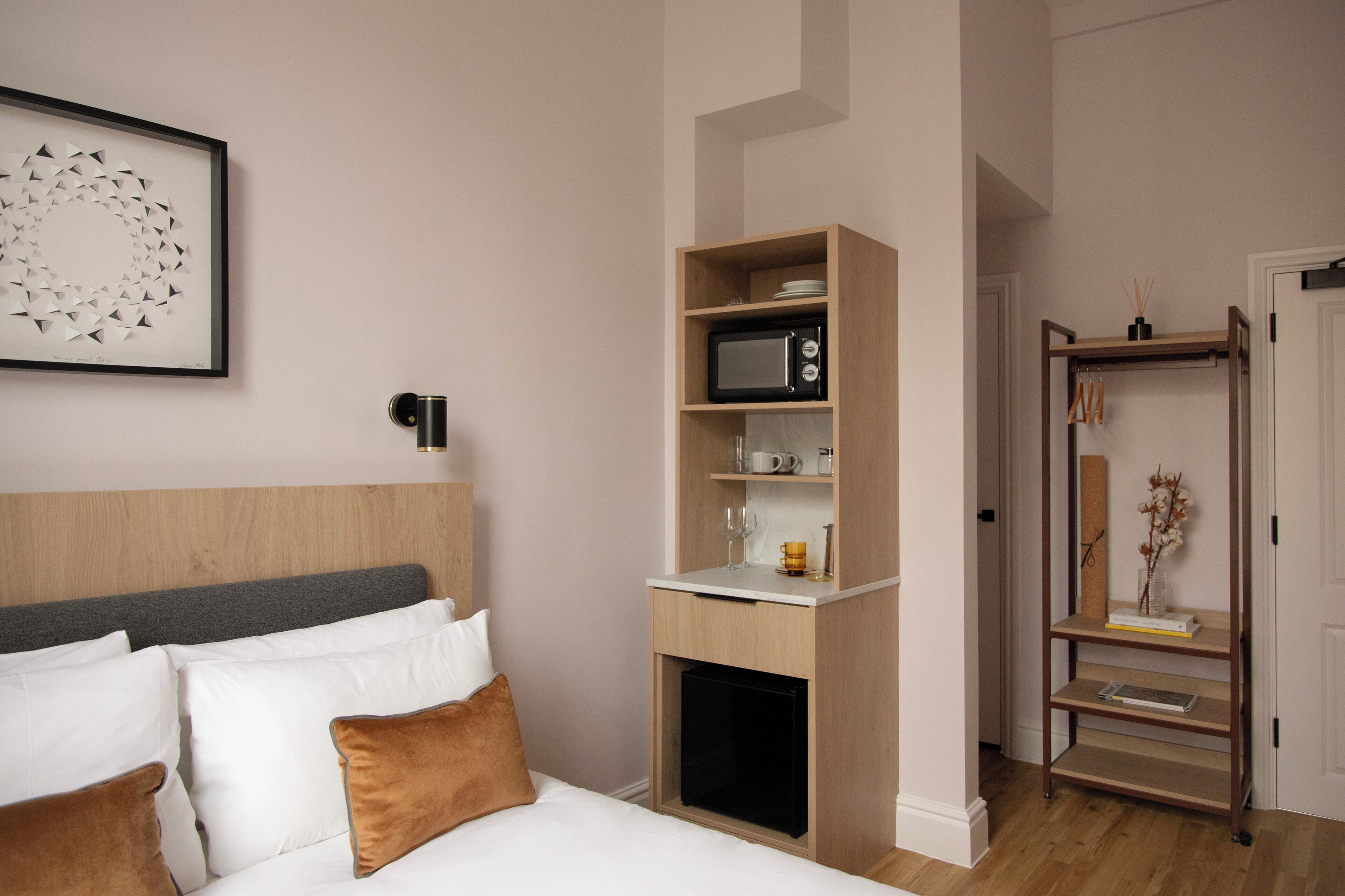 A hotel room with a bed, a bedside table with a microwave, a shelf with books and a vase, and a framed artwork on the wall.