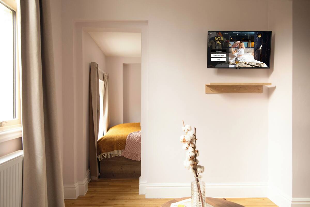 A bedroom with a TV mounted on the wall, a bedside table with a vase, and a window with curtains.