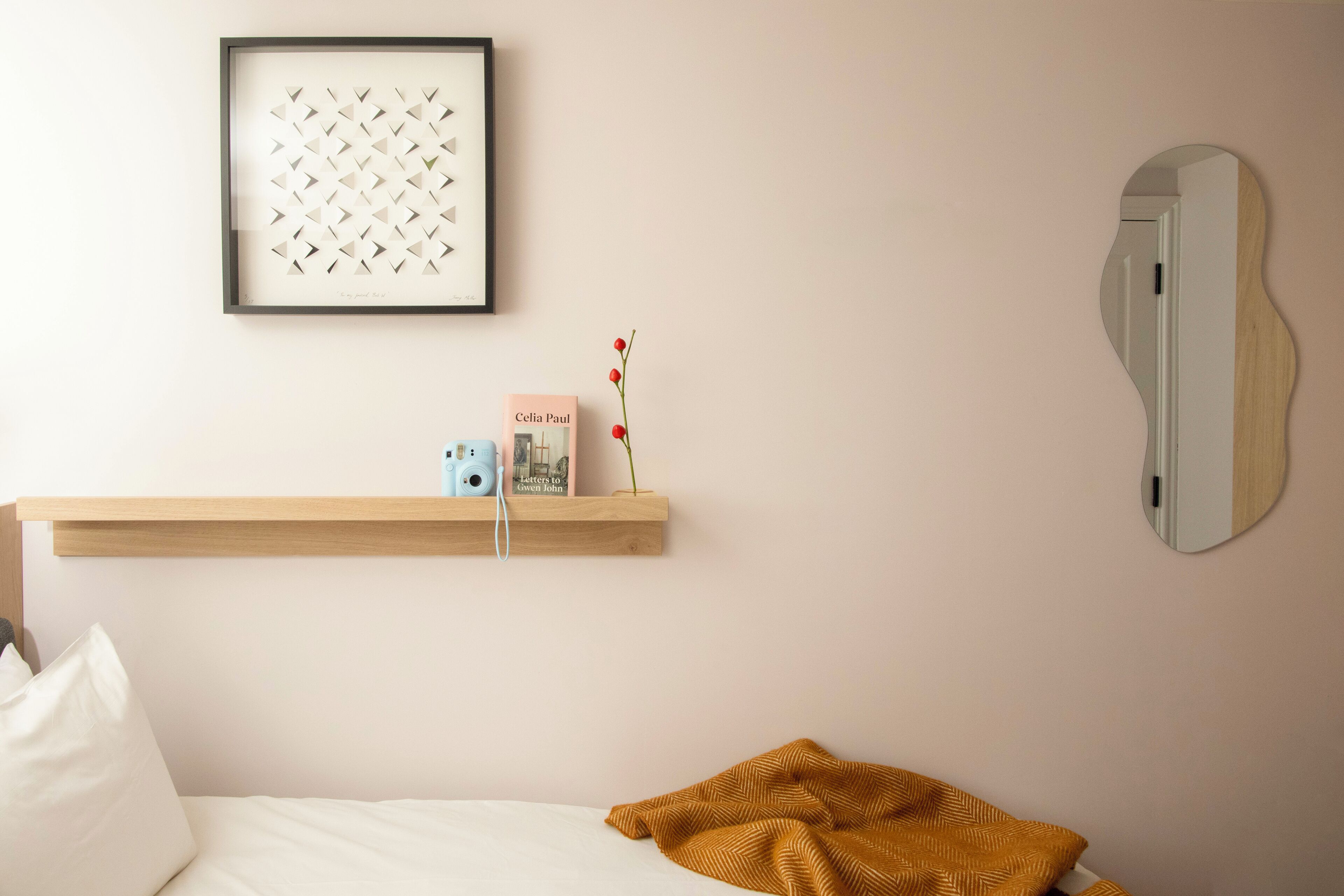 Une chambre à coucher avec un lit, une étagère en bois sur laquelle se trouvent un appareil photo et un livre, une œuvre d’art encadrée et un miroir.
