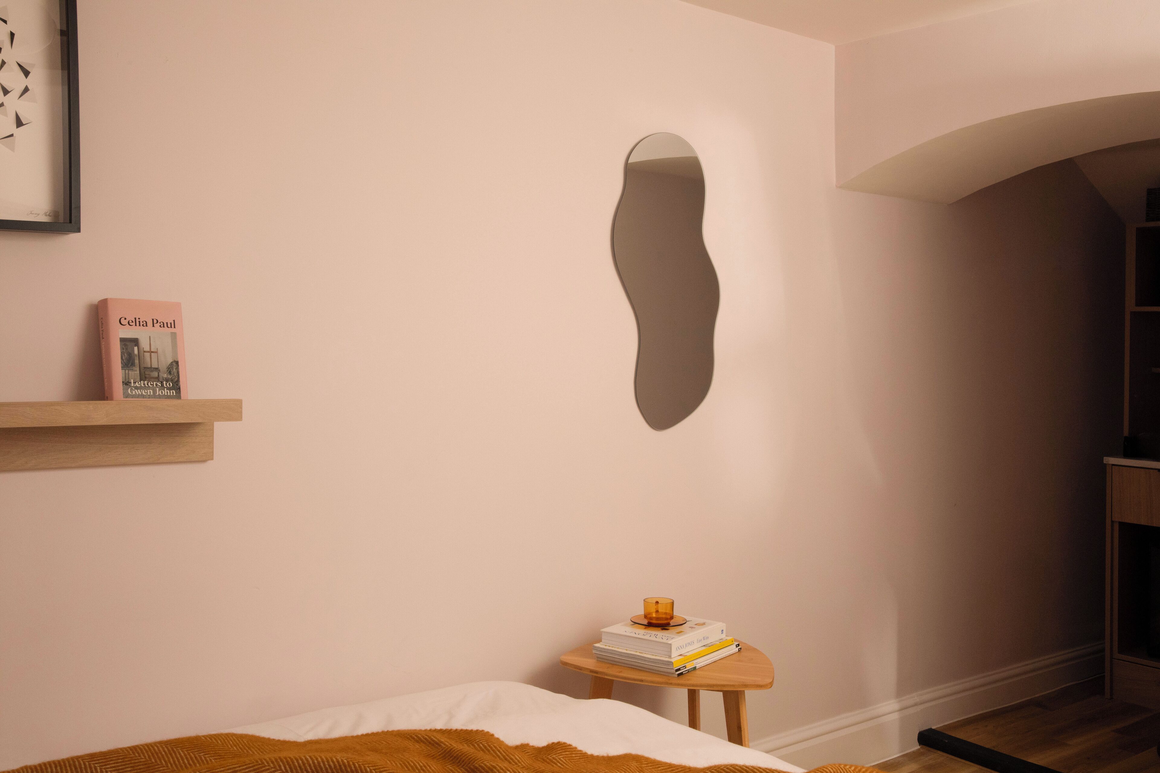 A bedroom with a bed, a bedside table with books and a candle, a framed picture, and a wall-mounted mirror.