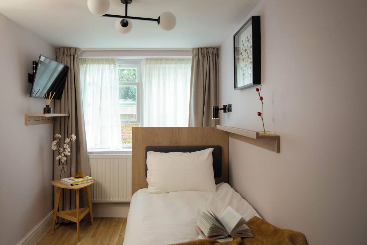 A bedroom with a bed, a bedside table with books, a TV mounted on the wall, and a window with curtains.