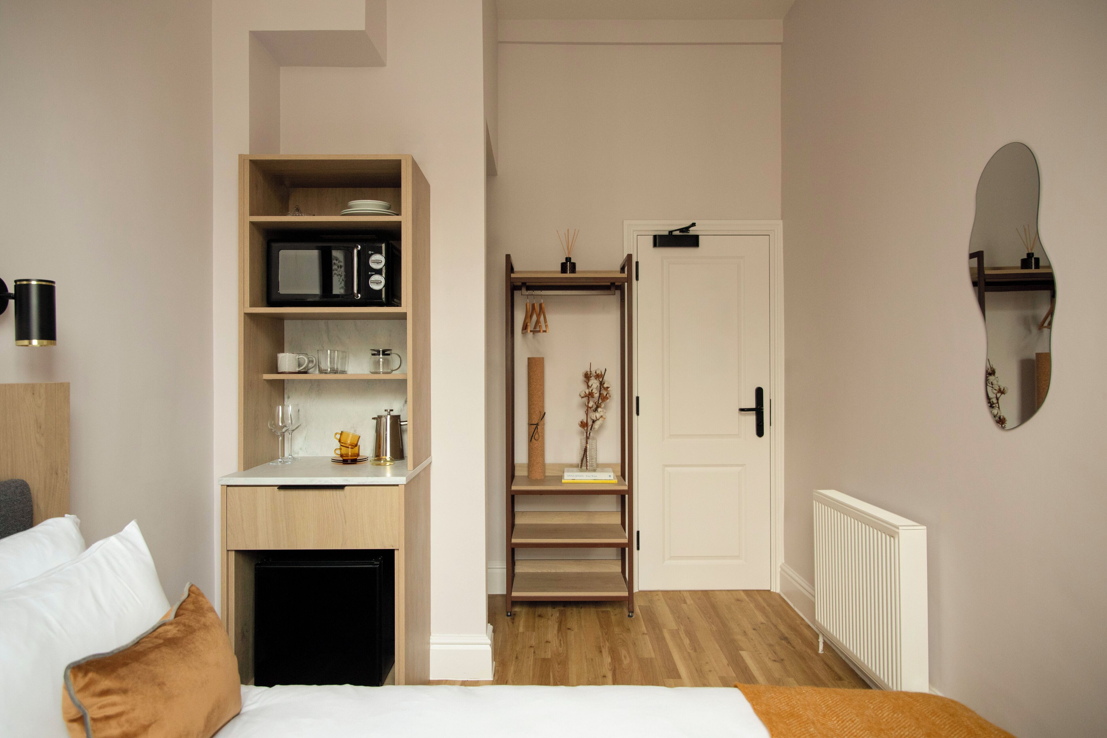A hotel room with a bed, a desk with a microwave, a bookshelf, a door leading to a staircase, and a radiator.
