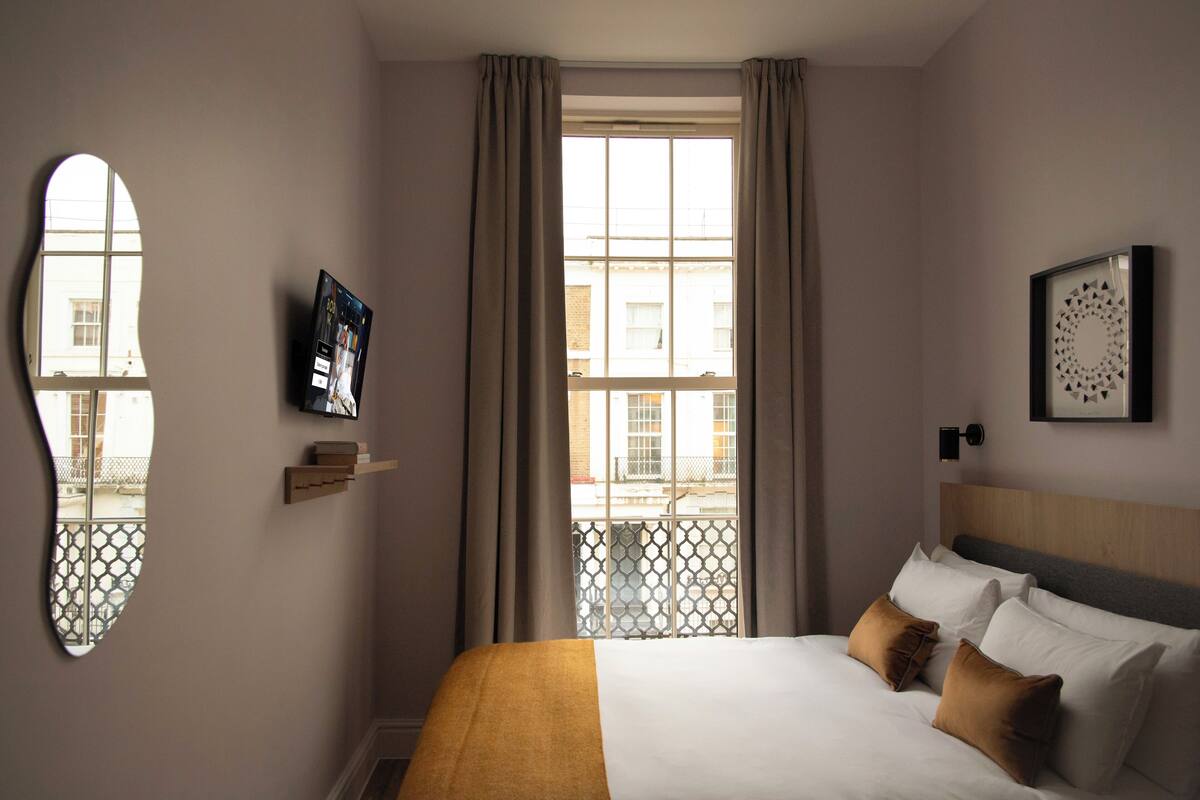 A hotel room with a large bed, a window with curtains, a mounted TV, and a wall-mounted artwork.