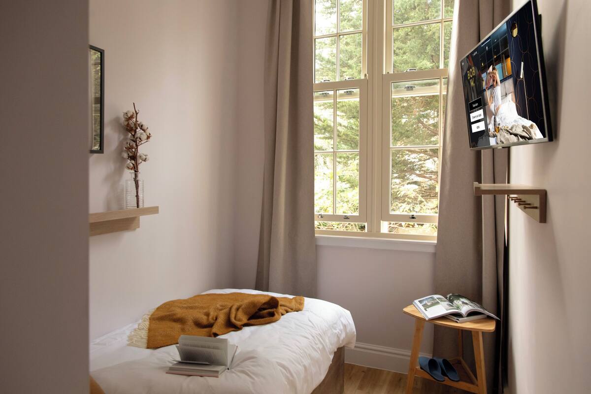 A bedroom with a bed, a laptop, a magazine, and a window with curtains.