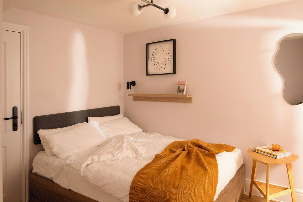 A neatly made bed with white linens and a brown throw blanket, a small wooden side table with a book and a cup, and a wall-mounted shelf with a framed picture and a small book.
