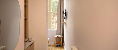 A narrow hallway with a wooden floor, a radiator, a window with curtains, and a small table with a book and slippers.