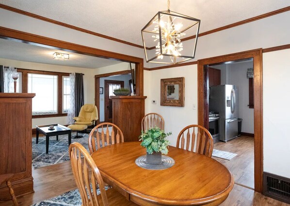 A dining area with a wooden table, chairs, and a chandelier. There is a kitchen visible in the background.