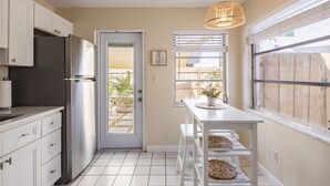 Una cocina con armarios blancos, un refrigerador de acero inoxidable, una ventana con persianas y una lámpara colgante.