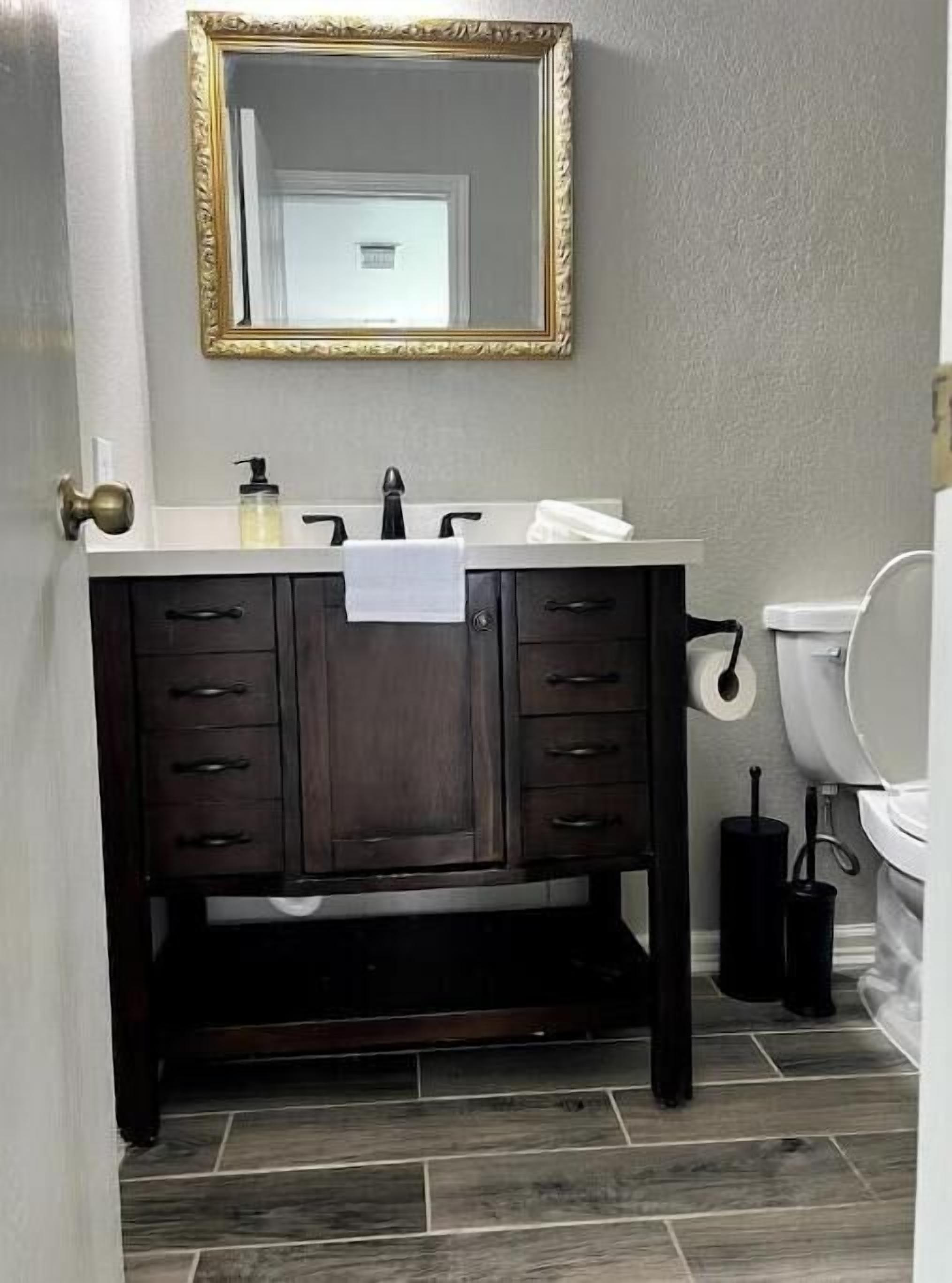 Ein Badezimmer mit einem dunklen Holz-Waschtisch, einem weißen Waschbecken, einem spiegel mit goldener Rahmung und einer Toilette.