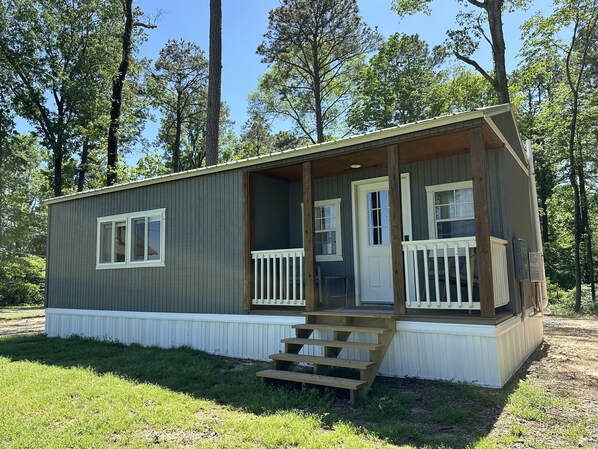 A small, single-story cabin with a porch, white railings, and a set of stairs leading to the entrance.