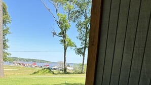 A porch with a white railing, a grey cushioned bench, and a view of a grassy field with trees and buildings in the distance. - D'Arbonne Nest Rentals: The Gator's Nest (Union Parish)