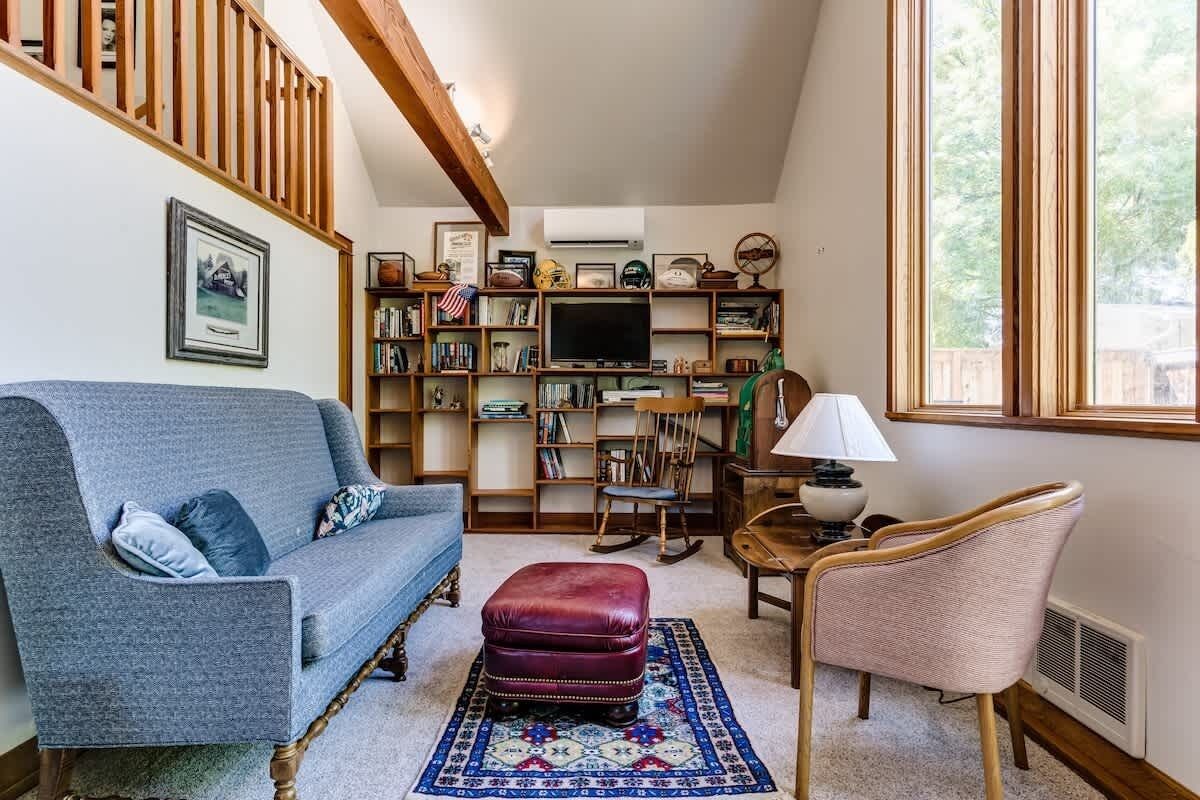 A cozy living room with a blue sofa, a red ottoman, a wooden rocking chair, a bookshelf, and a television.