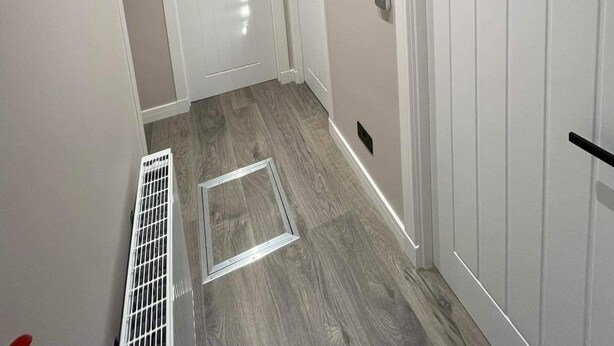 Un couloir avec une porte blanche, un miroir rond et une grille d’aération dans le sol.